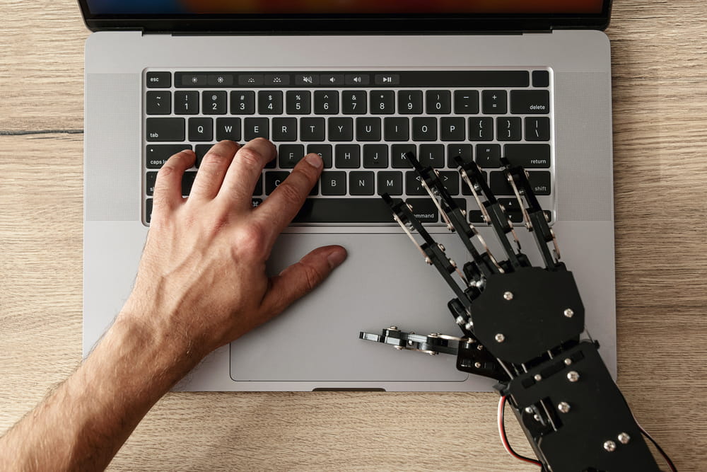 Human hand typing on a laptop keyboard beside a robotic hand